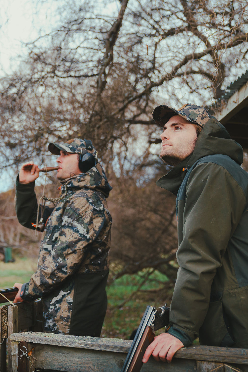 Two men in hunting gear standing outdoors with trees in the background