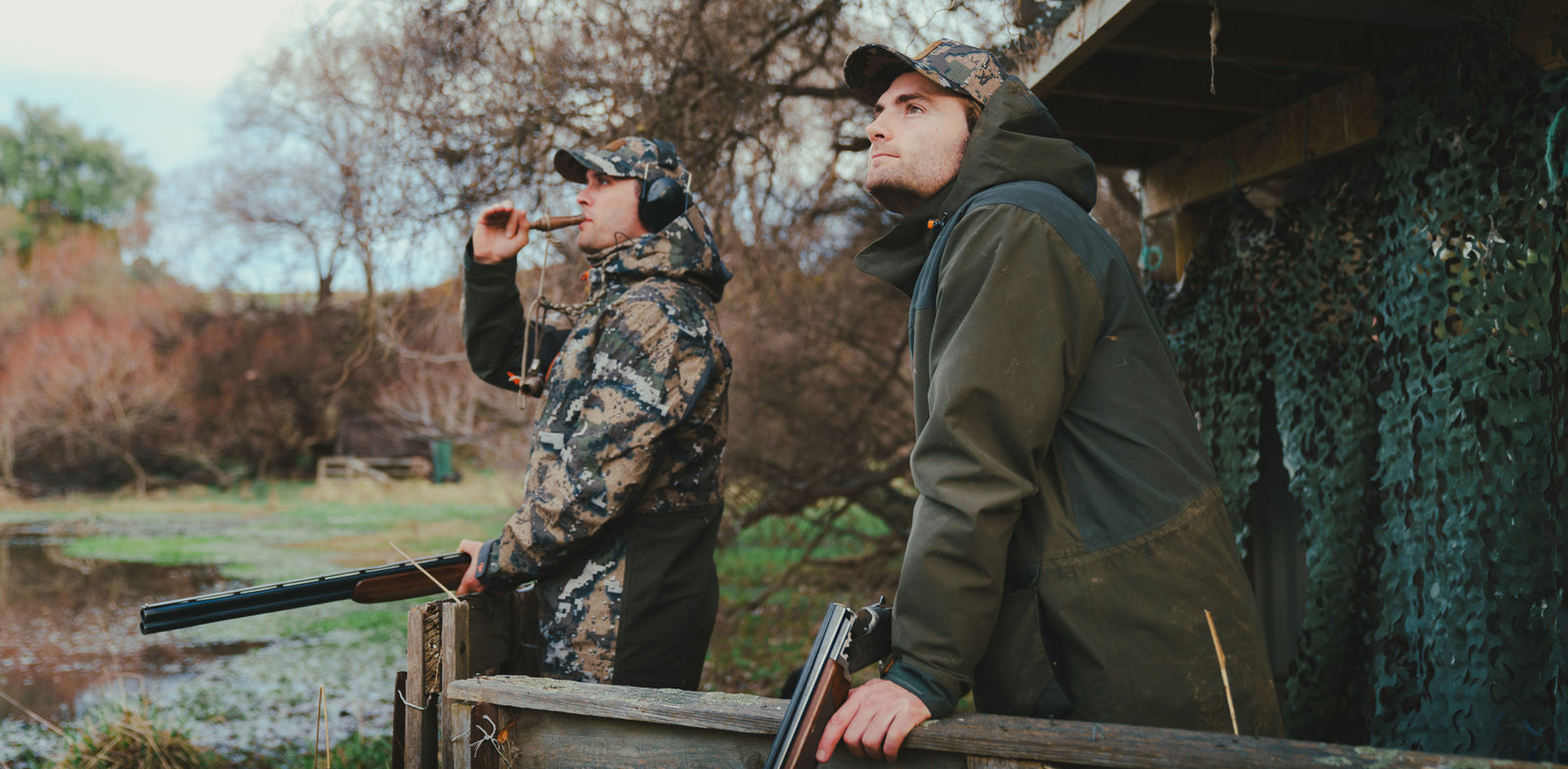 Two hunters in camouflage clothing standing near a wooden blind in a natural setting.