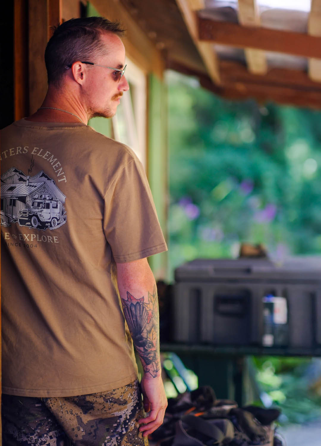 Man wearing a brown t-shirt with a graphic design, standing in a room with greenery outside.