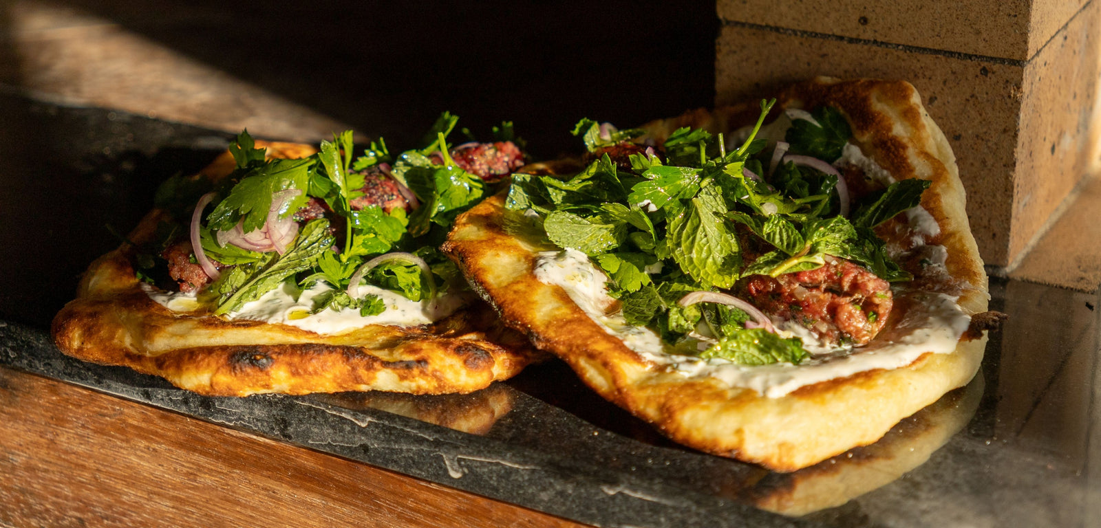 Two flatbreads with greens and other toppings on a stone surface.