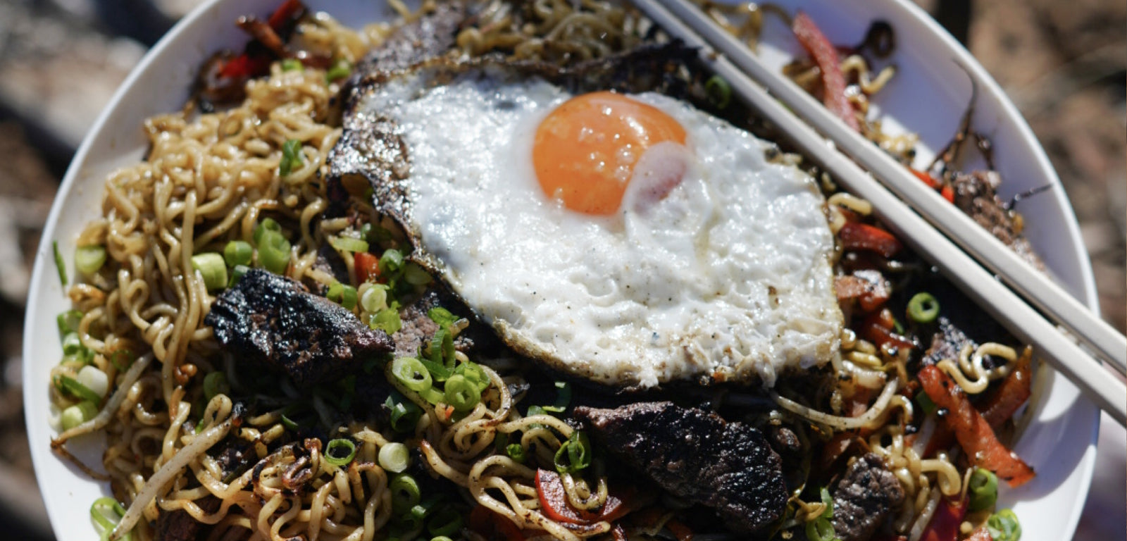 Noodles with a fried egg and vegetables on a white plate with chopsticks.